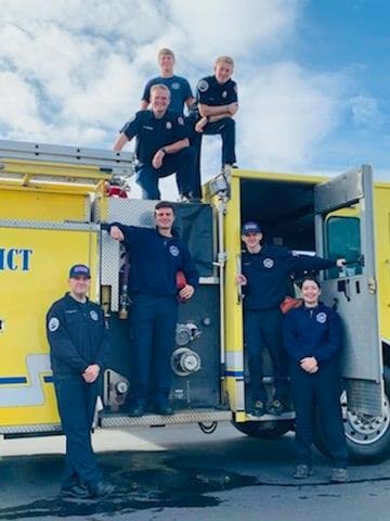 A group of people in uniforms posing on and around a yellow fire truck under a blue sky.