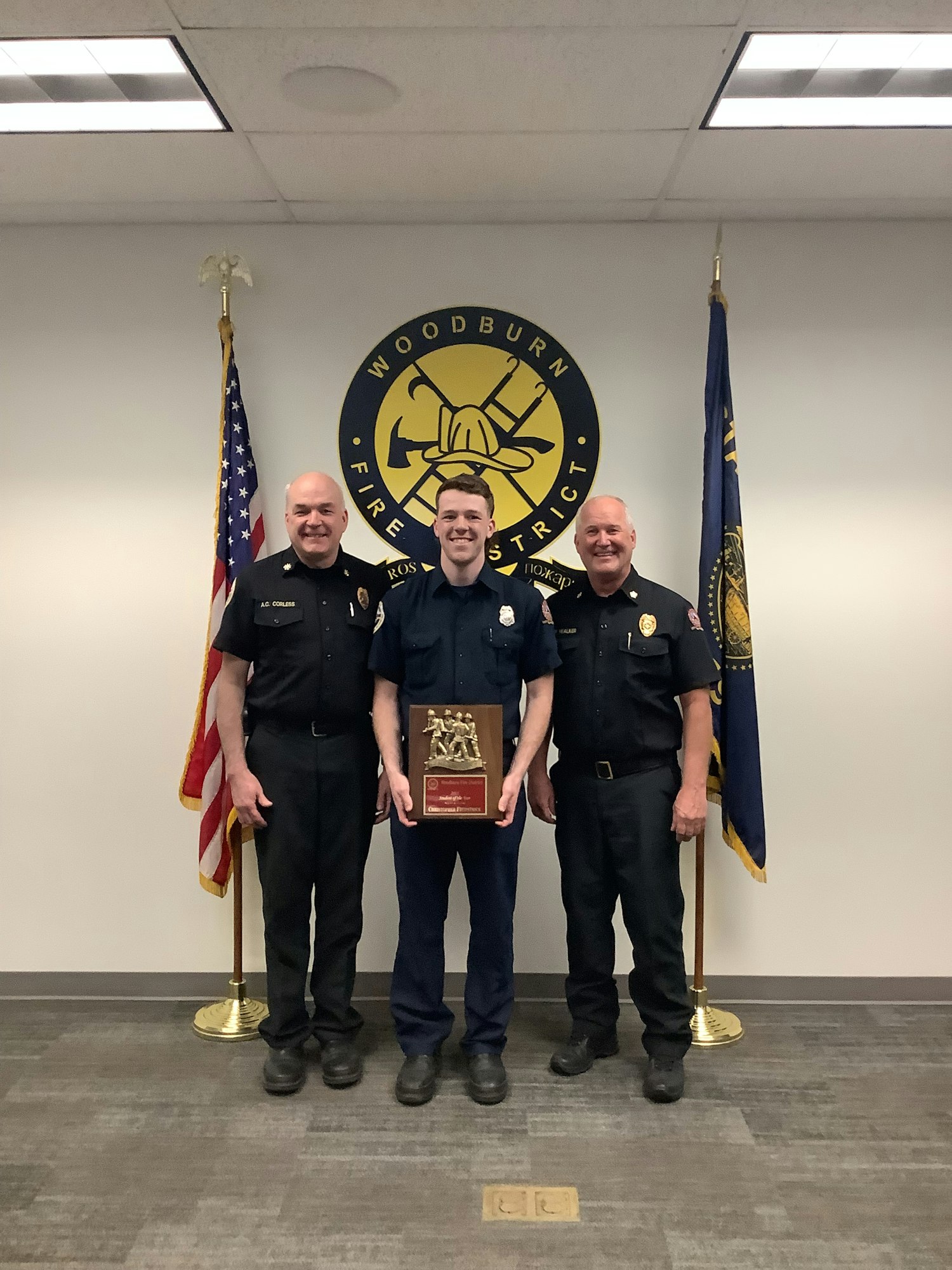 Three firefighters stand together in uniform, holding an award, in front of the Woodburn Fire District logo and flags.