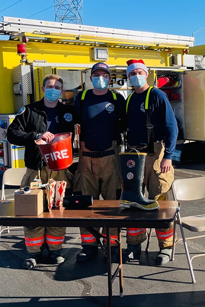Three firefighters in masks stand by a yellow fire truck, with a table of donation items, and a boot labeled "FIRE."