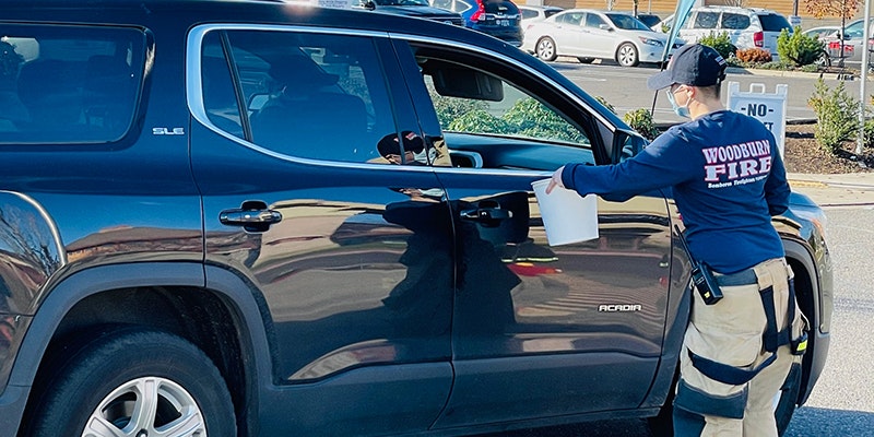 A person in a "Woodburn Fire" shirt hands a container to someone in a parked SUV.