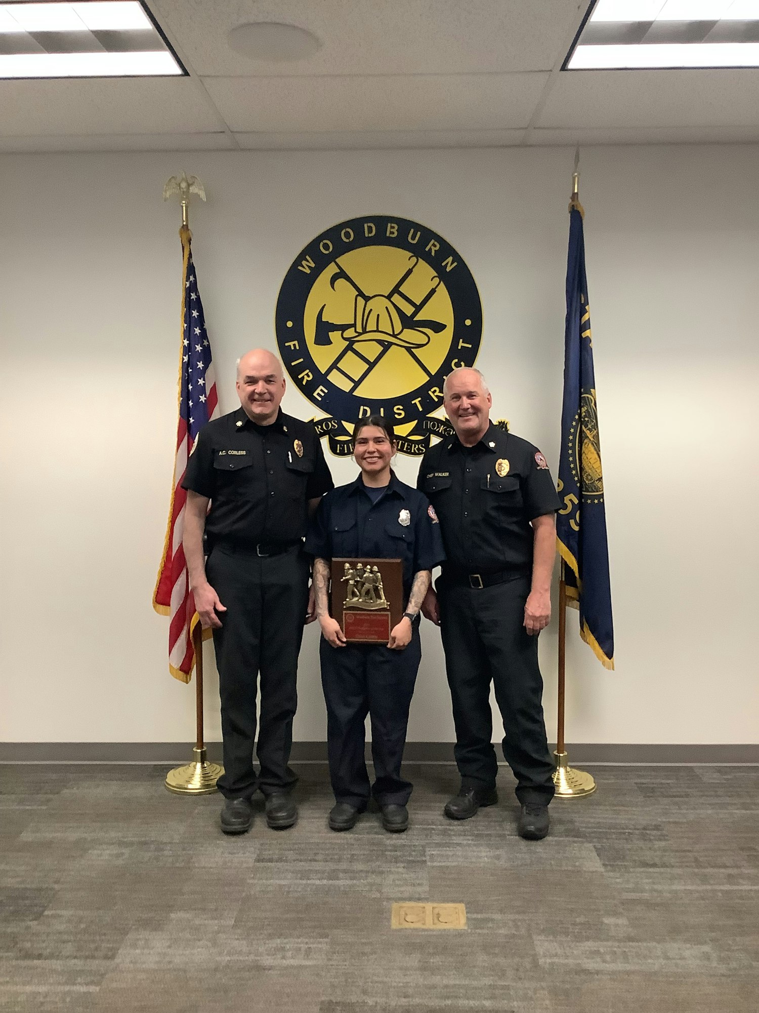 Three firefighters stand together holding an award, with flags and a fire district emblem in the background.