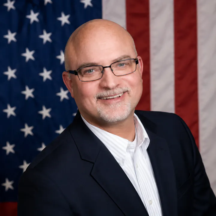 A smiling man in a dark suit and glasses poses in front of an American flag, showcasing a professional portrait style.