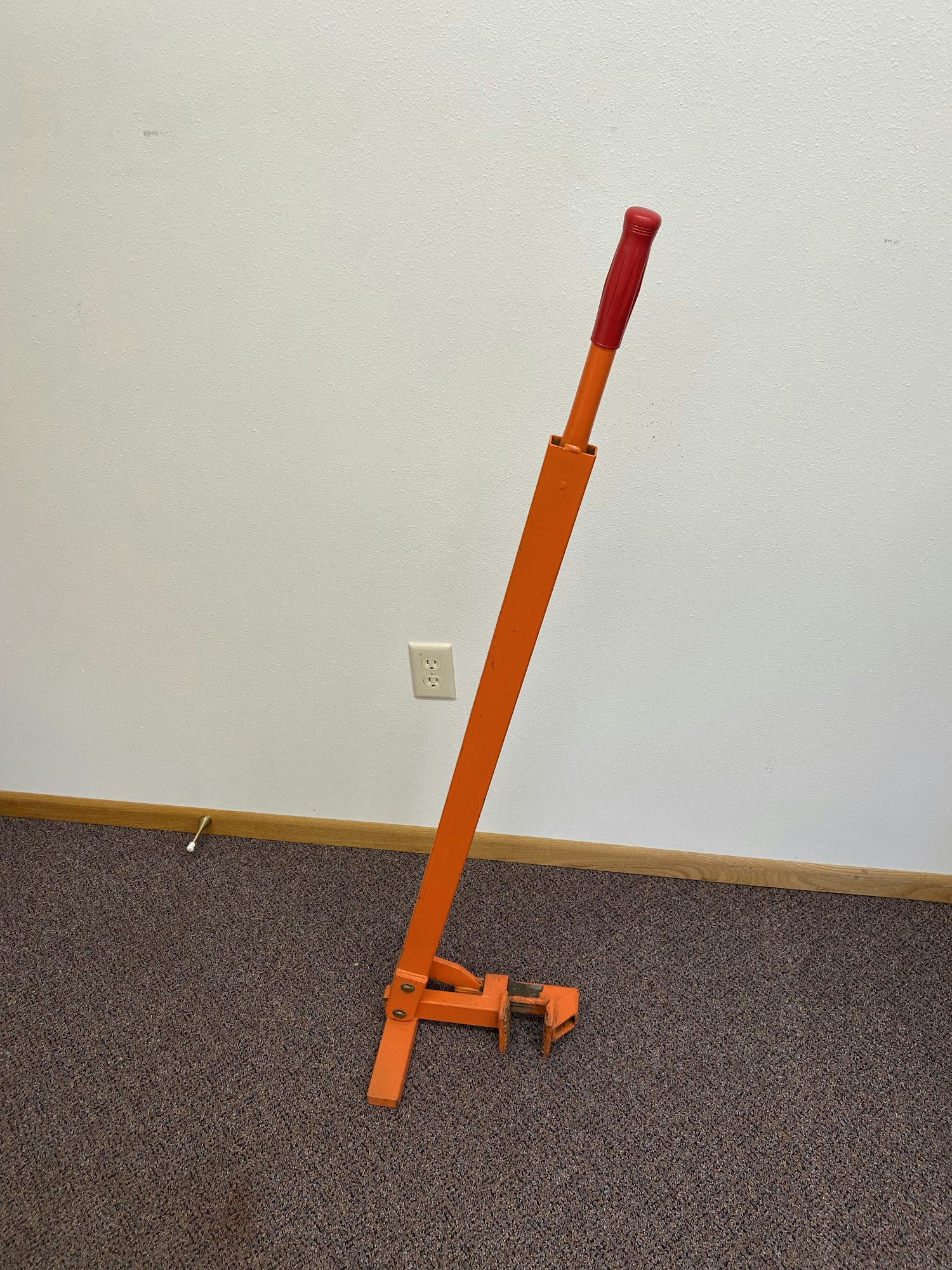 An orange carpet stretcher with a red handle leaning against a wall on a carpeted floor.