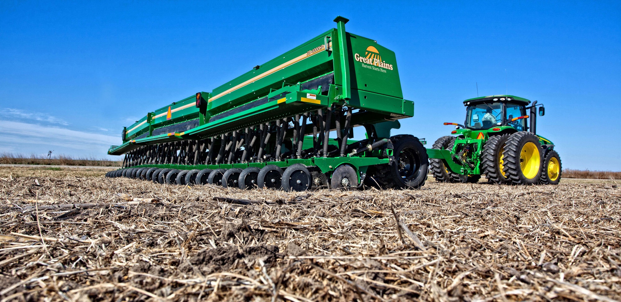 Tractor with a green no-till seed in a dry field covered in crop residue under a clear blue sky.