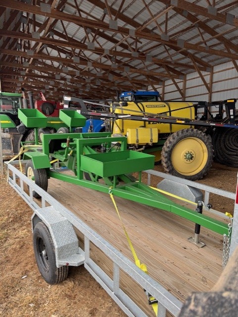 The image shows a green agricultural machine mounted on a trailer inside a barn, with other machinery visible in the background.