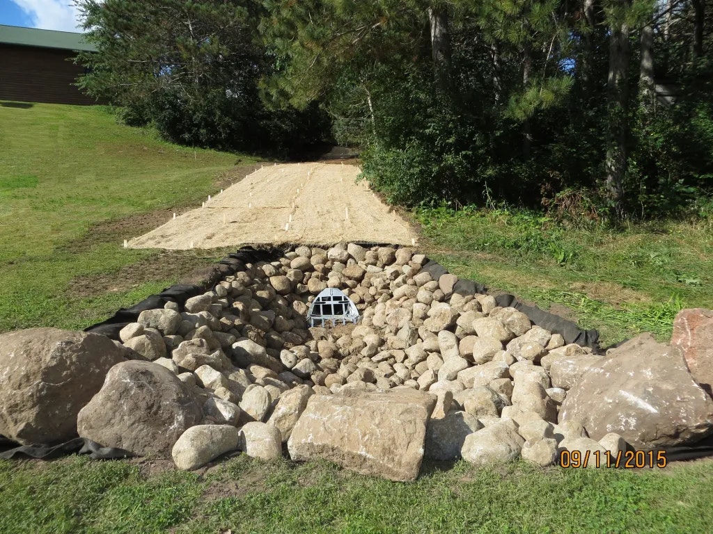 A riprap drainage outlet with rocks for erosion control, leading from a fabric-covered slope.