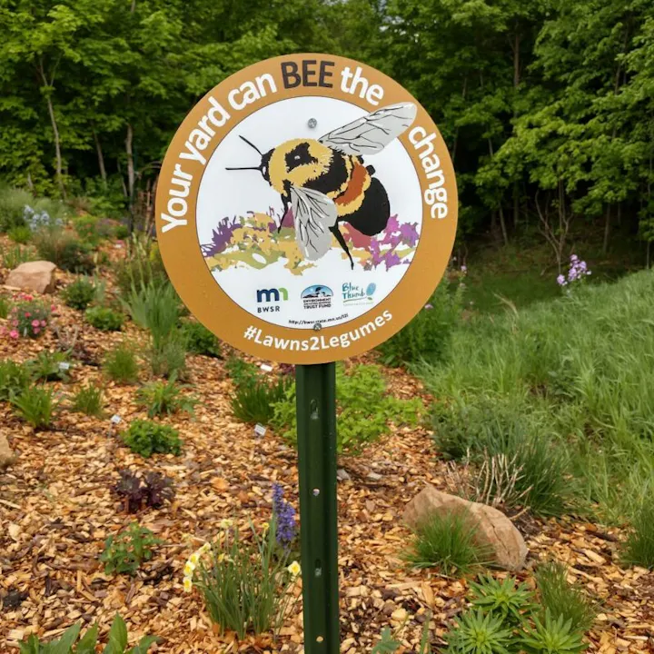 A sign with a bee illustration reads "Your yard can BEE the change" and #Lawns2Legumes, in a garden setting.