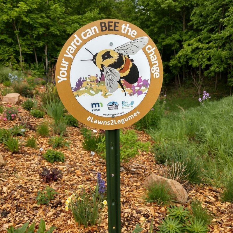 A sign with a bee illustration reads "Your yard can BEE the change" and #Lawns2Legumes, in a garden setting.