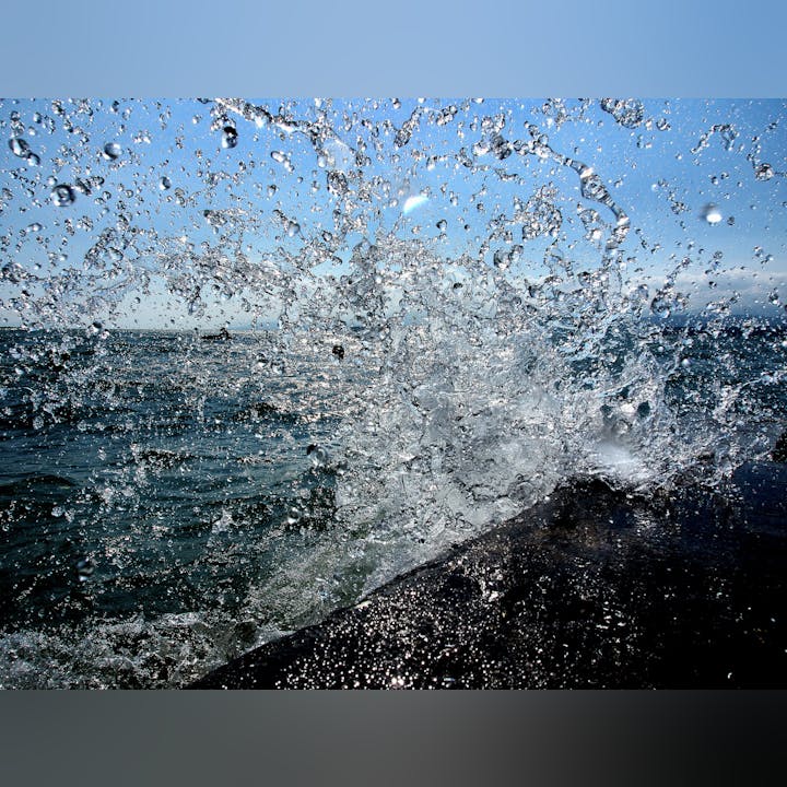 Ocean waves crashing against rocks, creating a spray of water droplets under a clear blue sky.