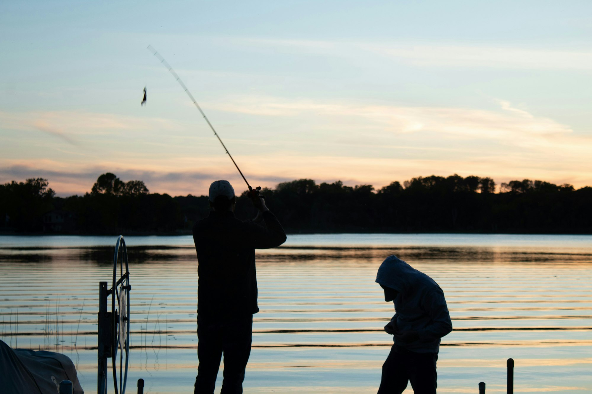 Two people fishing at sunset by a calm lake with trees in the background.