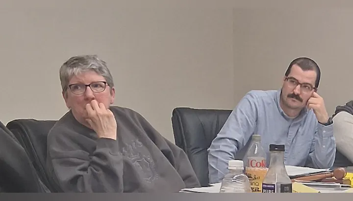 Two people sitting at a table, deep in thought during a meeting, with drinks and a gavel on the table.