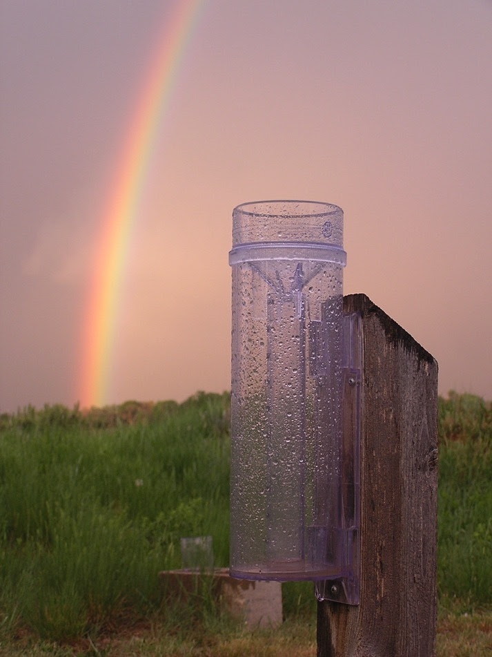 A rain gauge on a wooden post with a rainbow in the background over a grassy field.