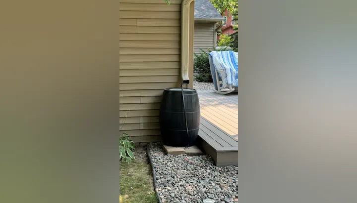 A rain barrel connected to a gutter downspout beside a house, near a deck and some rocks.