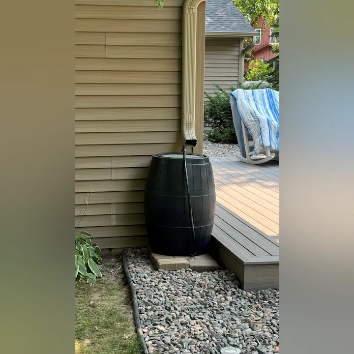 A rain barrel connected to a gutter downspout beside a house, near a deck and some rocks.