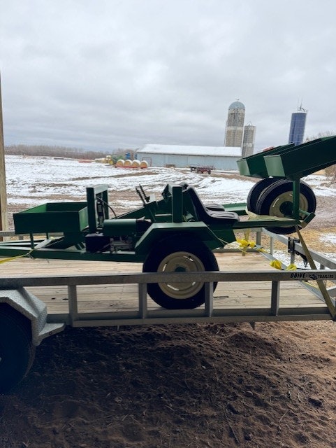 The image shows a green agricultural trailer or equipment loaded onto a trailer, with a snowy outdoor backdrop.
