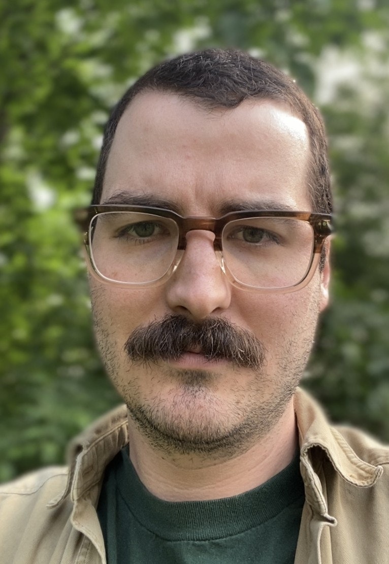 A person with glasses and a mustache outdoors, wearing a green shirt and beige jacket.