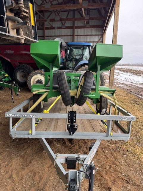 Image features a green agricultural implement on a utility trailer, with a tractor in the background, set in a barn environment.