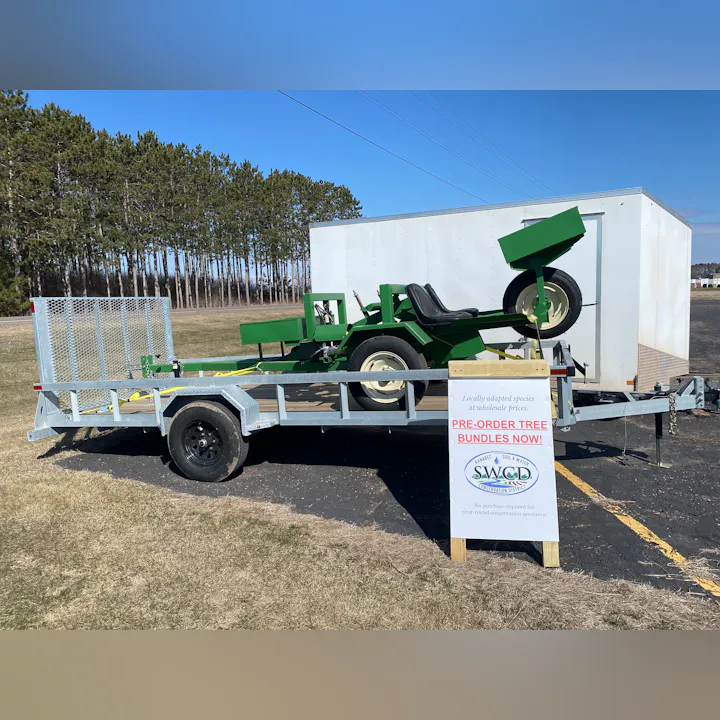 A green equipment trailer with a sign promoting pre-orders for tree bundles, set against a backdrop of trees.