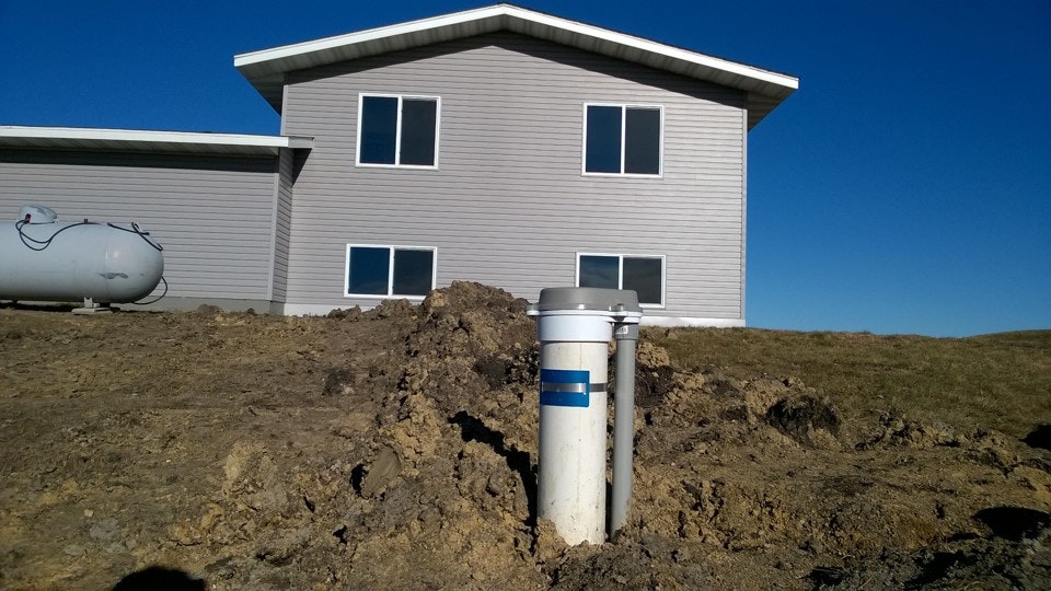 A house with a propane tank and two pipes sticking out of the ground in front, set against a clear blue sky.
