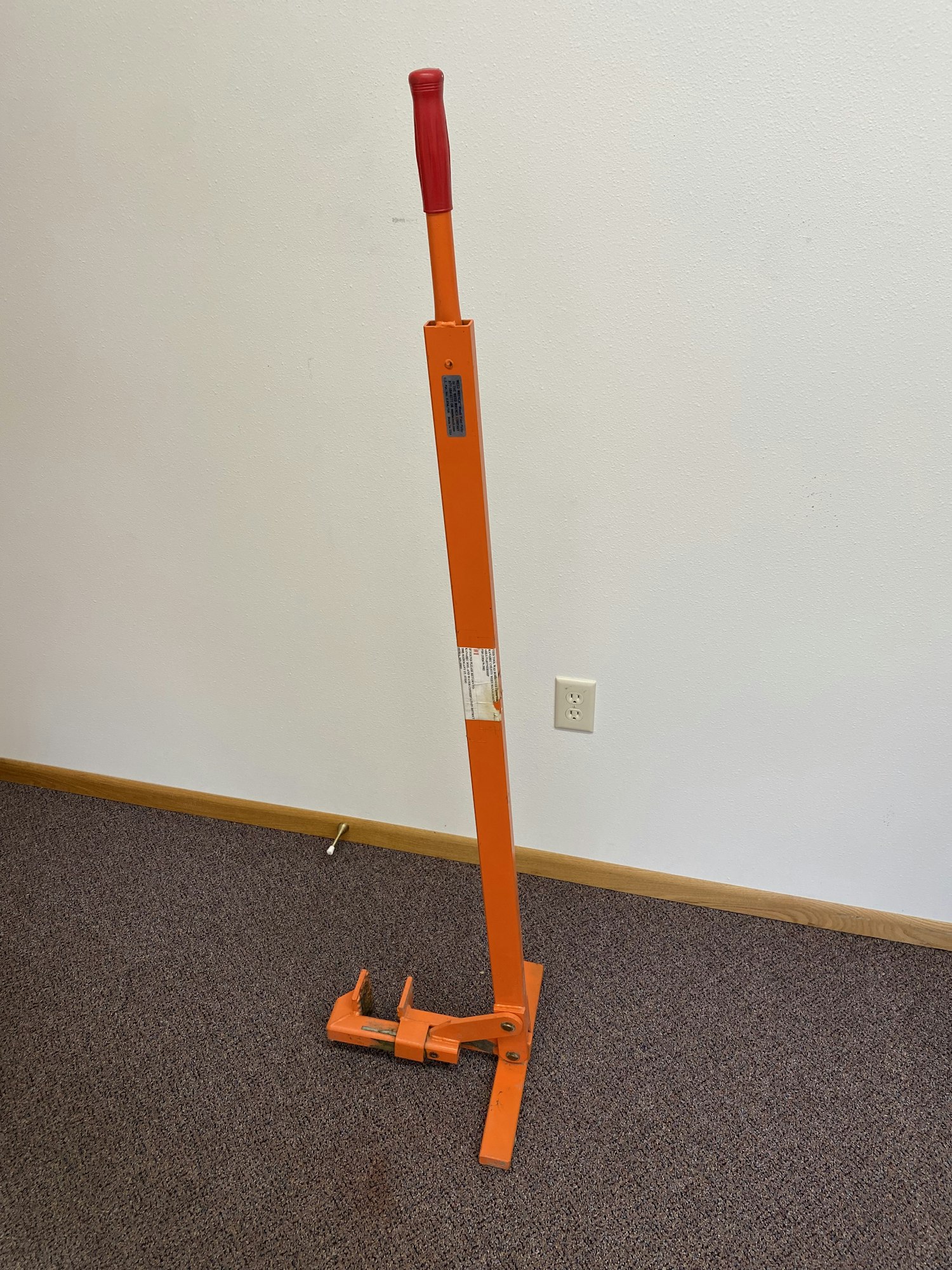 An orange carpet stretcher with a long handle leaning against a carpeted floor and white wall.