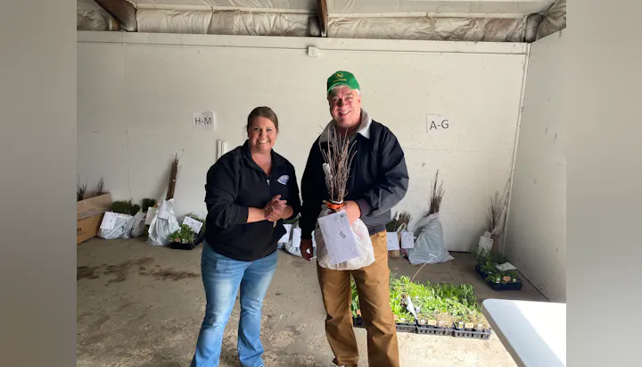A man and woman pose together in a storage area with plants and gardening materials, smiling and holding a bundle of sticks.