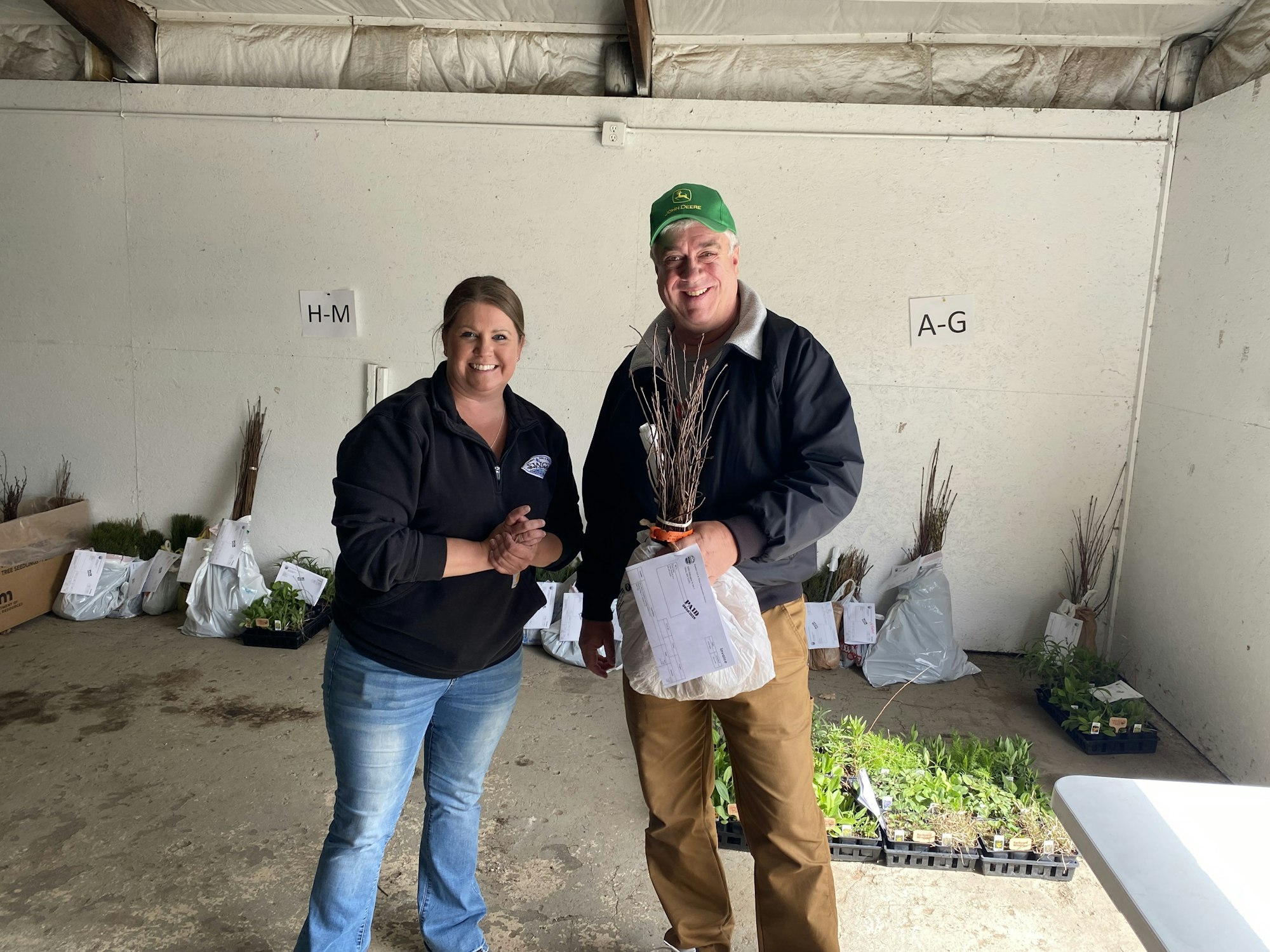 A man and woman pose together in a storage area with plants and gardening materials, smiling and holding a bundle of sticks.