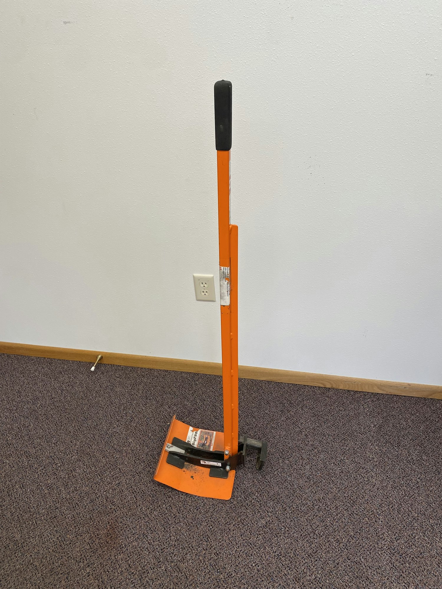 An orange tile floor scraper with a black handle is leaning against a wall.