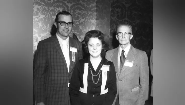 Three people dressed formally, wearing name tags, stand in front of a patterned wallpaper.