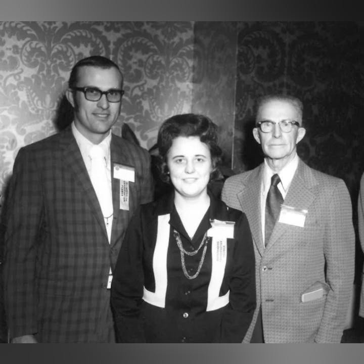 Three people dressed formally, wearing name tags, stand in front of a patterned wallpaper.