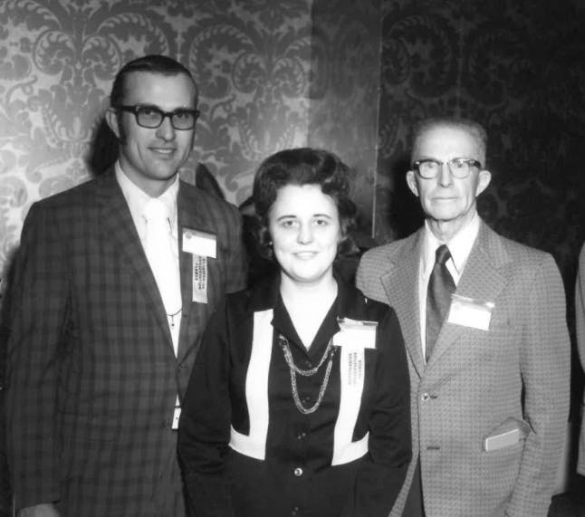 Three people dressed formally, wearing name tags, stand in front of a patterned wallpaper.