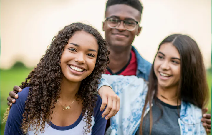 Three smiling young people stand together outdoors, enjoying a friendly moment with a green background.