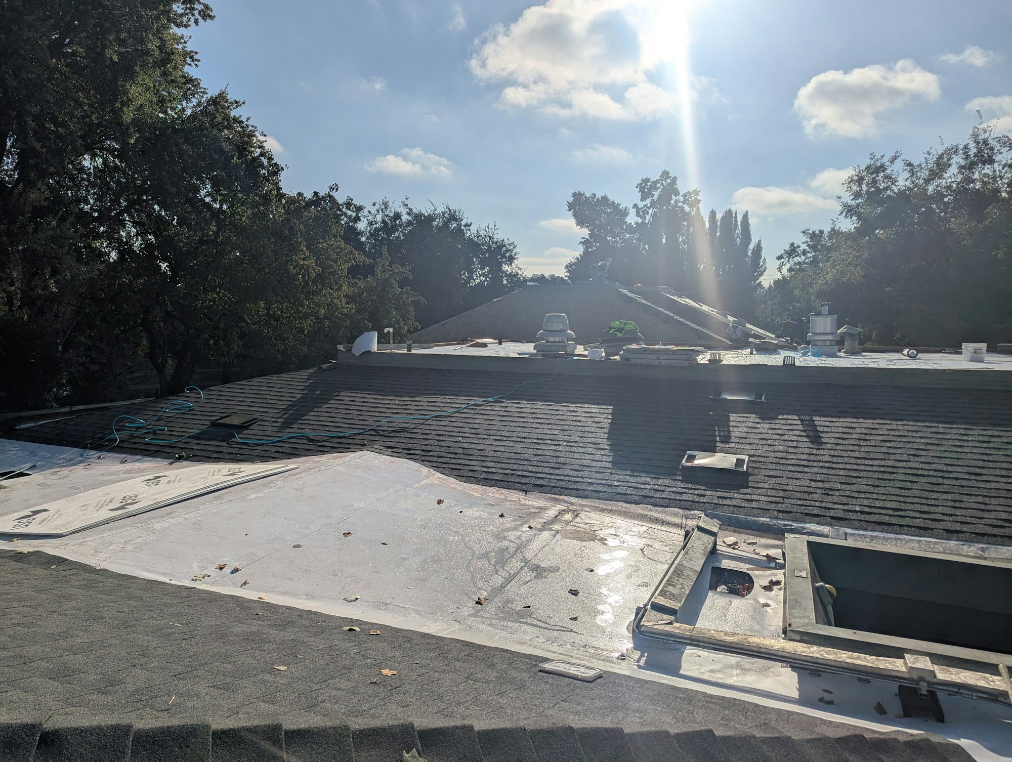 Rooftop under renovation with shingles, tools, and materials; surrounded by trees, with the sun shining in the background.