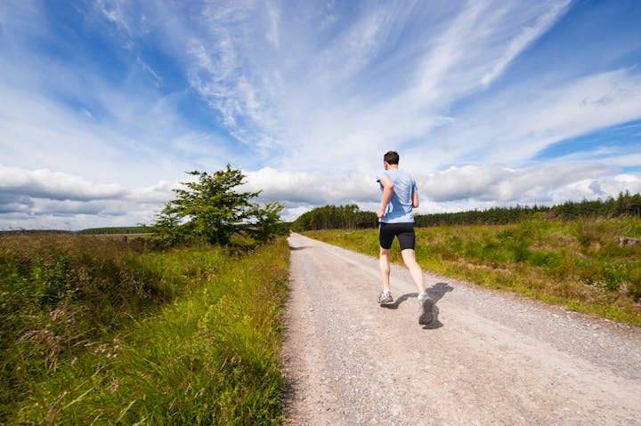 A person running on a trail with grass on both sides under a blue sky with clouds.