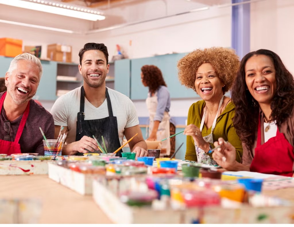 A group of four people, wearing aprons, smiling and painting together in an art studio setting.