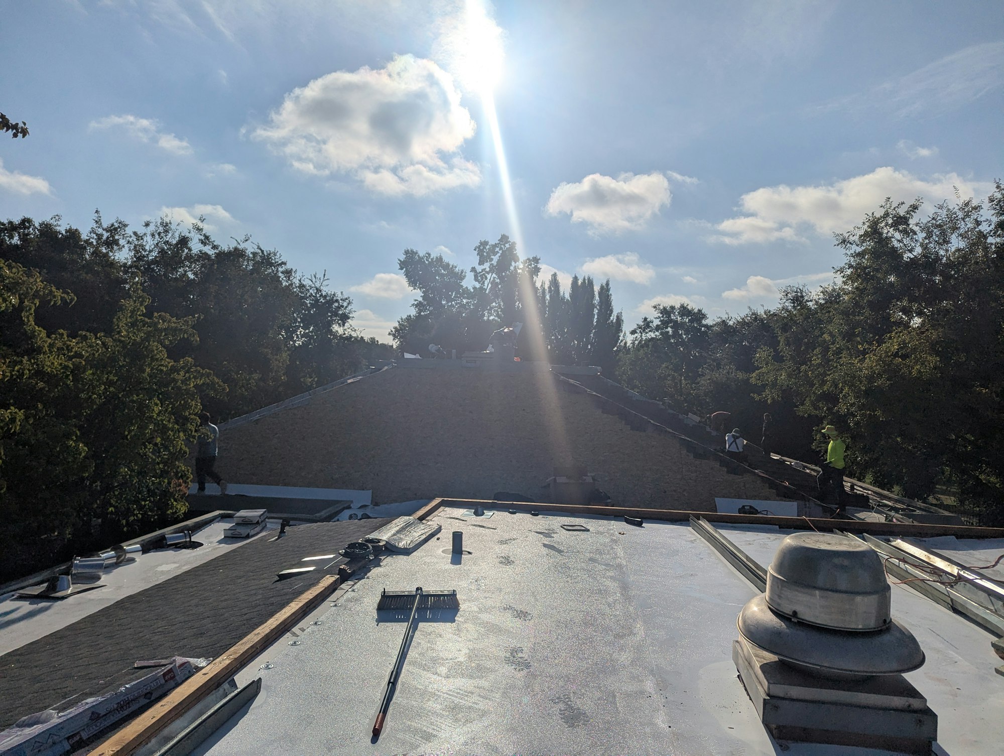 Workers on a roof under the sun, with tools and materials visible, surrounded by trees and a clear sky with clouds.