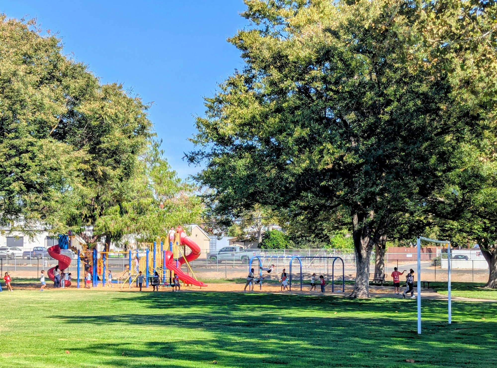 A sunny park scene with children playing on a colorful playground and swings under large trees.