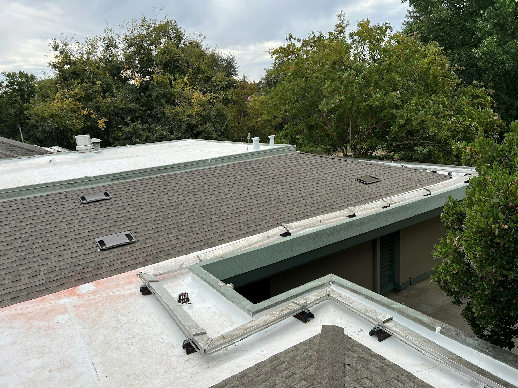 Flat and sloped roofs with shingles and vents, surrounded by trees.