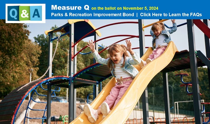 Two kids sliding down a slide in a playground, with a sign about a parks bond measure.