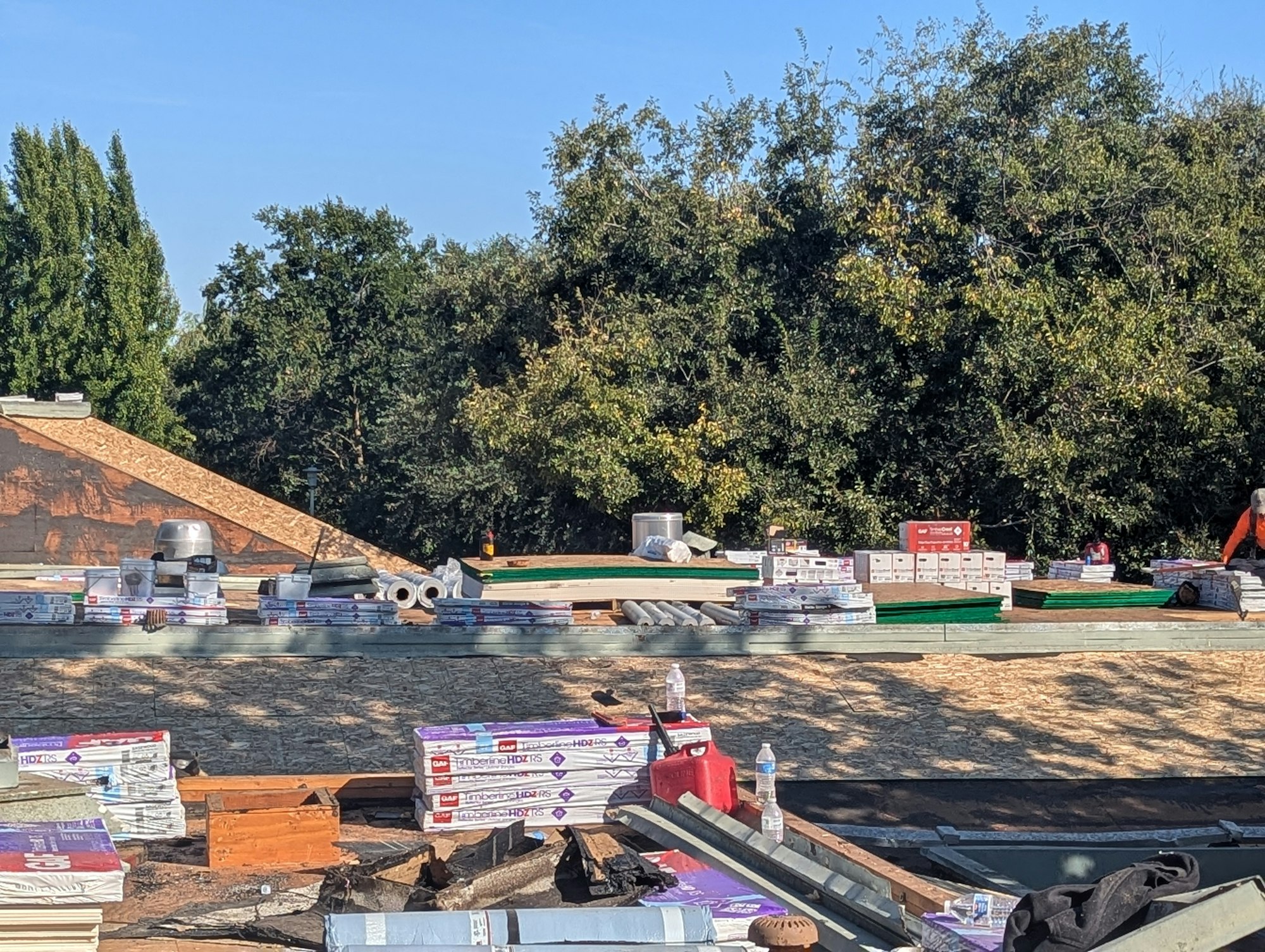 Roof under construction with materials like wood, shingles, and insulation. Worker in orange visible; trees in background.
