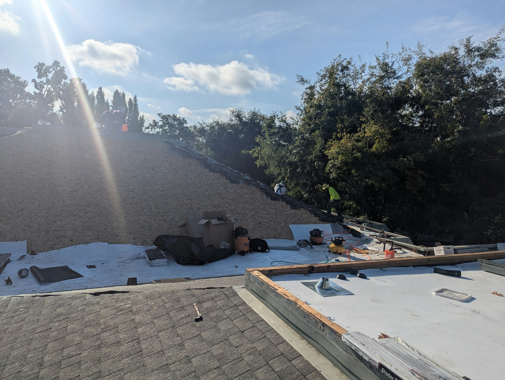 Construction workers are installing a roof, with tools and materials scattered on a partially covered structure. Trees and sky in the background.
