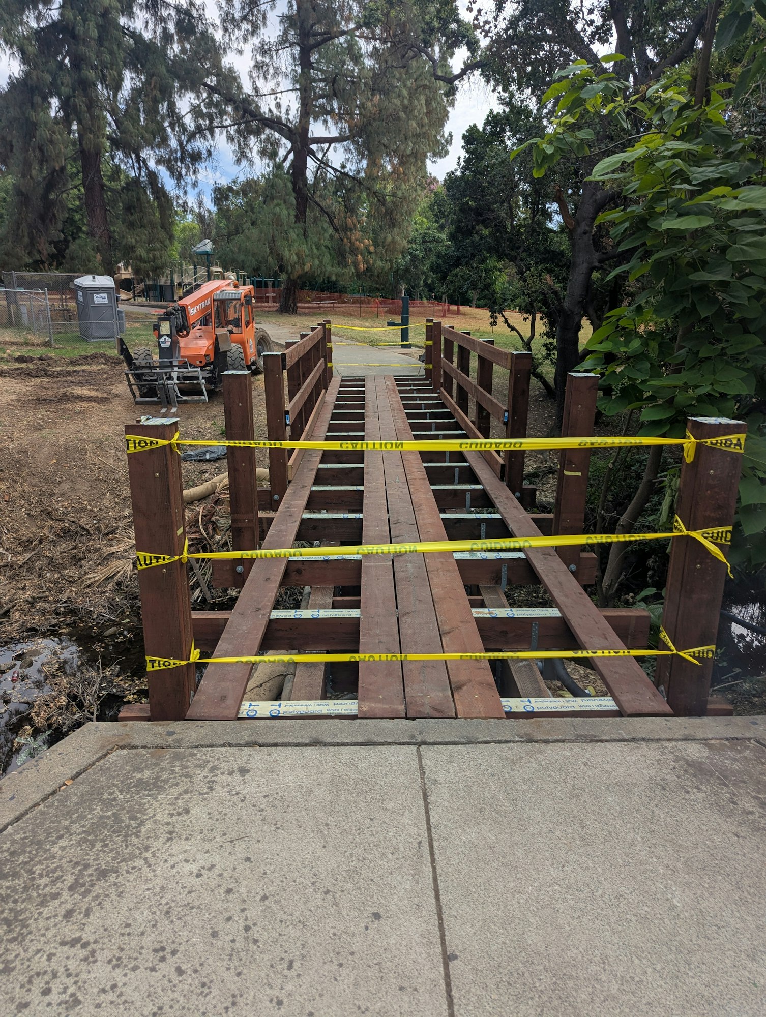 A wooden bridge under construction or repair, marked with caution tape, near trees and construction equipment in a park.