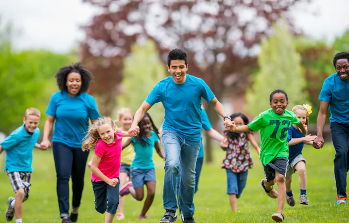A group of children and adults in matching blue shirts happily run together in a sunny park setting.
