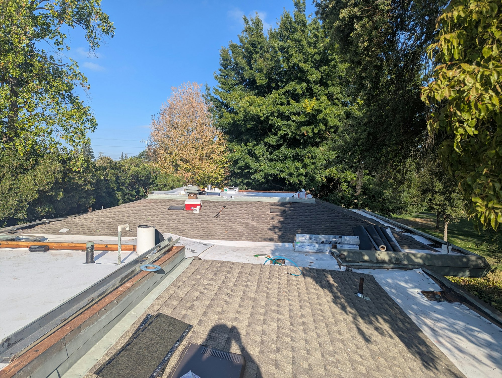 A rooftop with vents and construction materials, surrounded by trees under a clear blue sky.