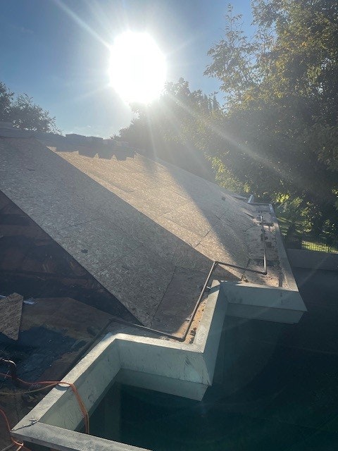A sunlit construction scene with a partially completed roof, surrounded by trees, under a bright sun.