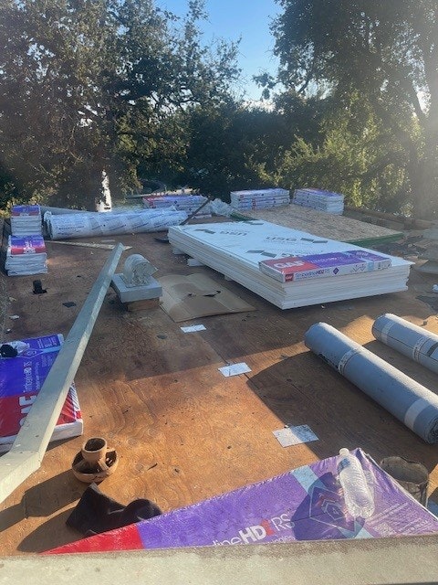 A rooftop with roofing materials, including underlayment rolls, insulation panels, and shingles, under trees and clear sky.