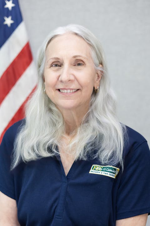 A woman with long gray hair in a navy shirt is standing in front of a U.S. flag.