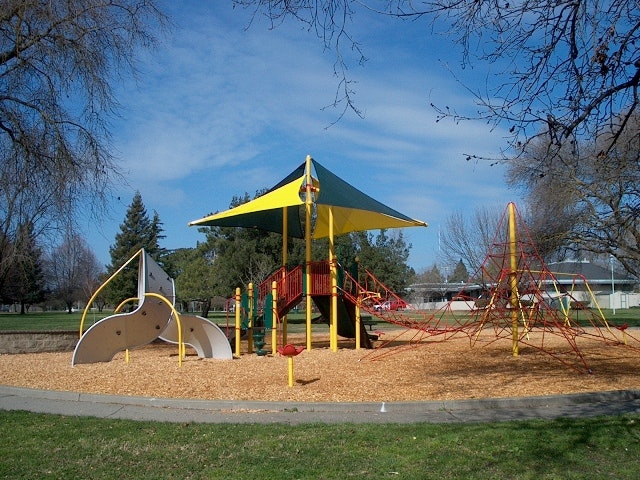 A colorful playground featuring climbing structures, a shaded area, and wood chip flooring set in a park with trees and blue sky.