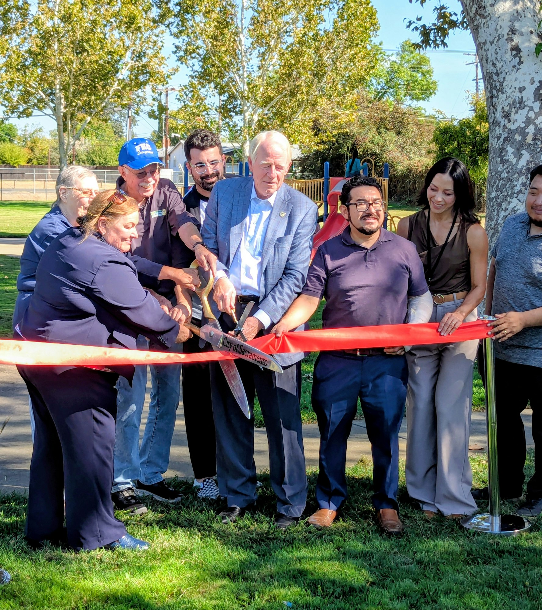 A group of people is gathered for a ribbon-cutting ceremony, celebrating the opening of a new park or facility.
