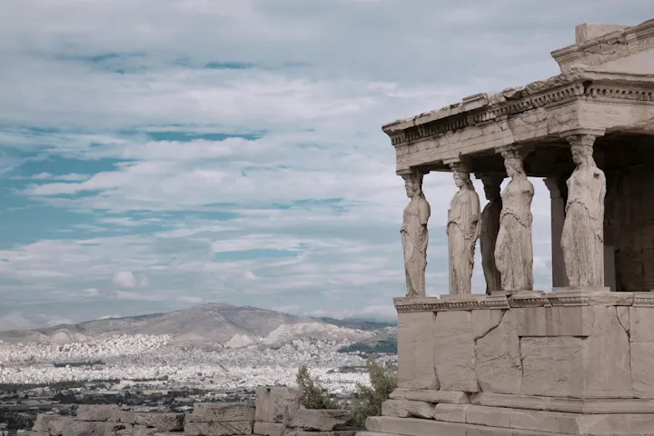 The image shows the Erechtheion in Athens, featuring Caryatids and a panoramic view of the city below against a cloudy sky.