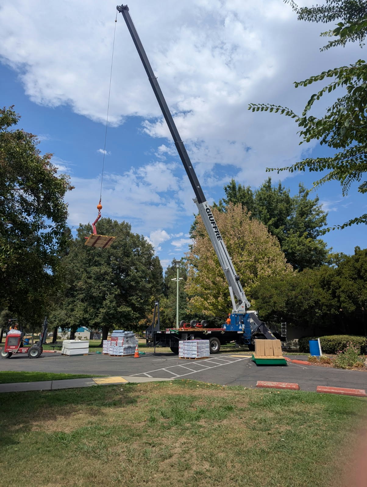 A crane lifts a pallet near a truck with building materials in a parking lot, surrounded by trees under a partly cloudy sky.